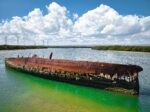 A drone shot of a ship wreck in green blueish water, surrounded by little pockets of Islands, blue sky covered with fluffy clouds