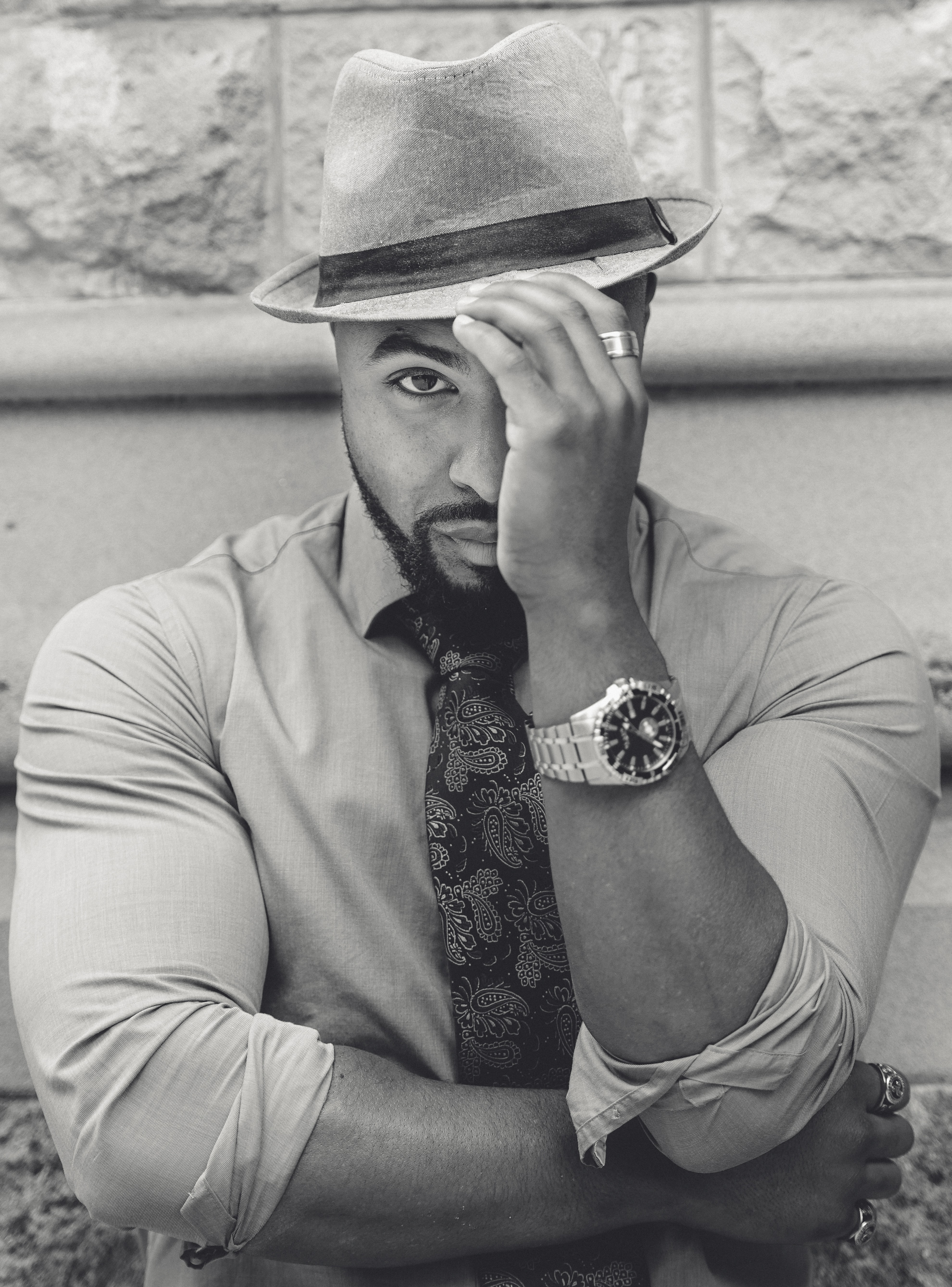 Black and white photo of Benjamin wearing a hat and standing against a wall with one folded arm.